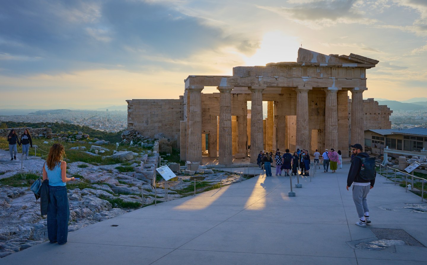 Turisti esplorano antiche rovine di un tempio al tramonto con la città in lontananza.